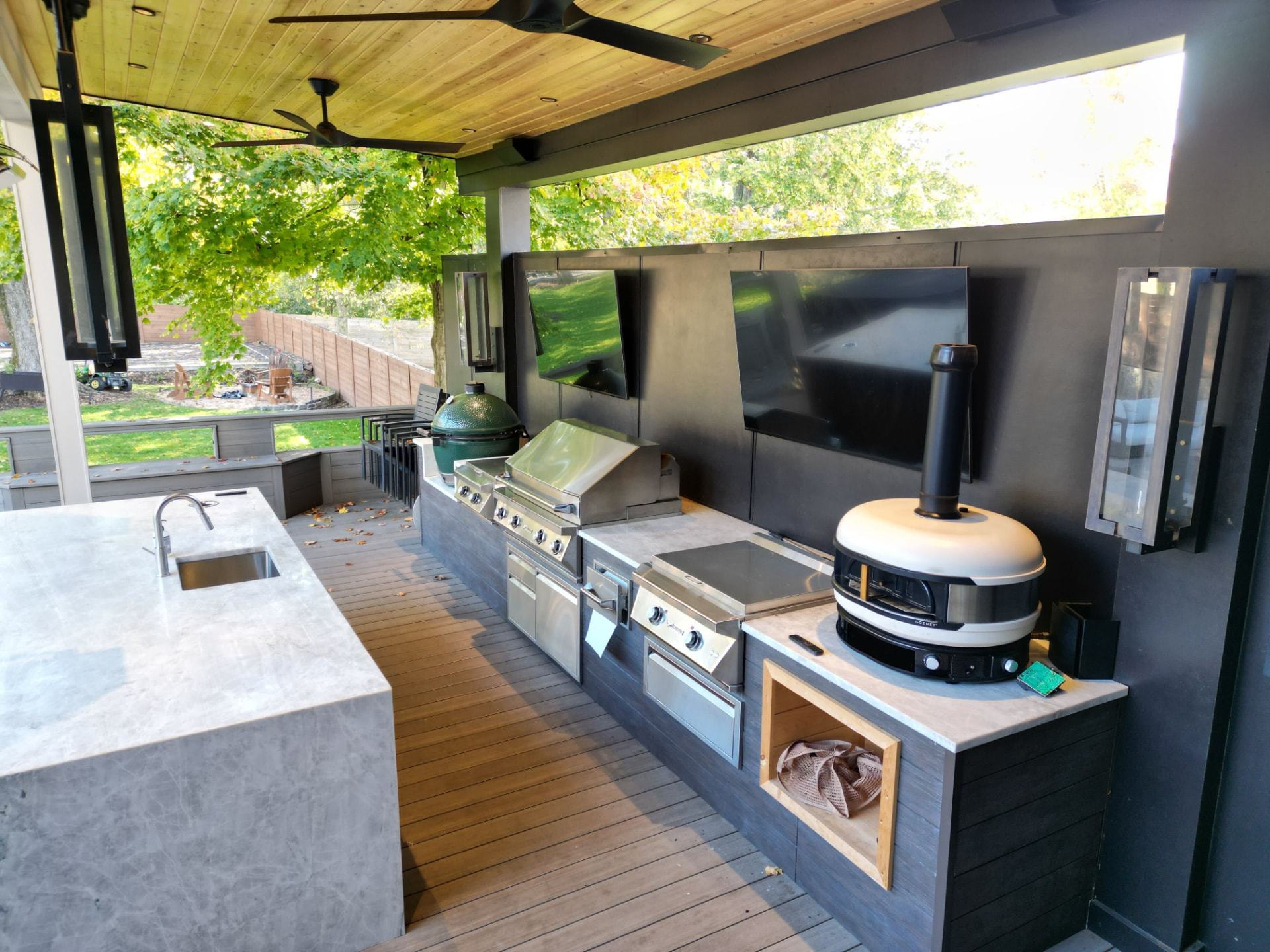 Covered outdoor kitchen with concrete island, grill, pizza oven and mounted televisions